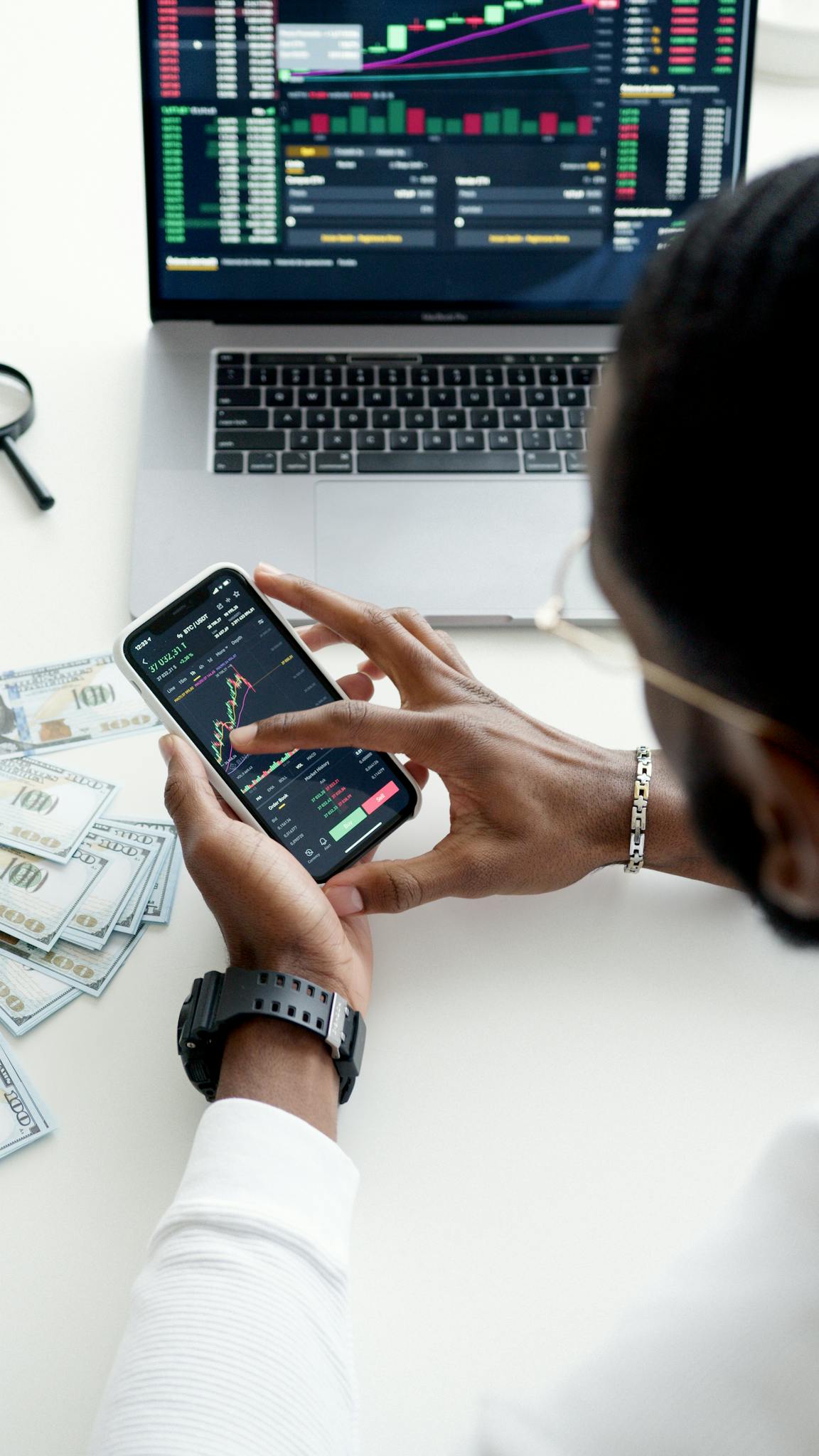 A person trading stocks using a smartphone, showcasing financial data on mobile and laptop screens.