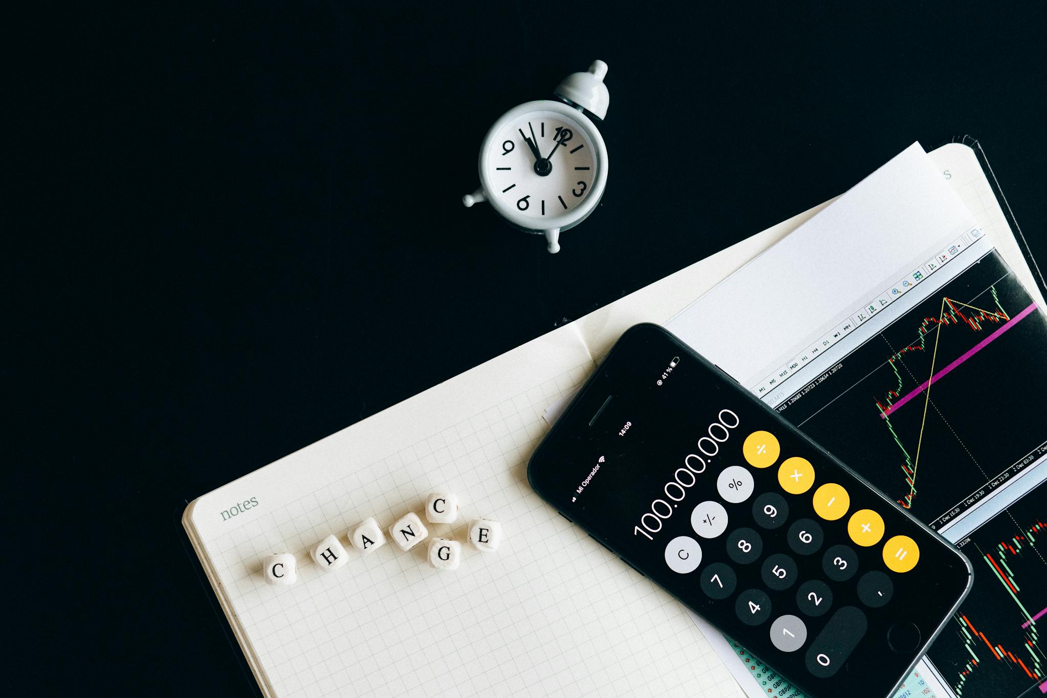 Notebook with an iPhone displaying 100,000,000, desk clock and financial charts suggesting market analysis.