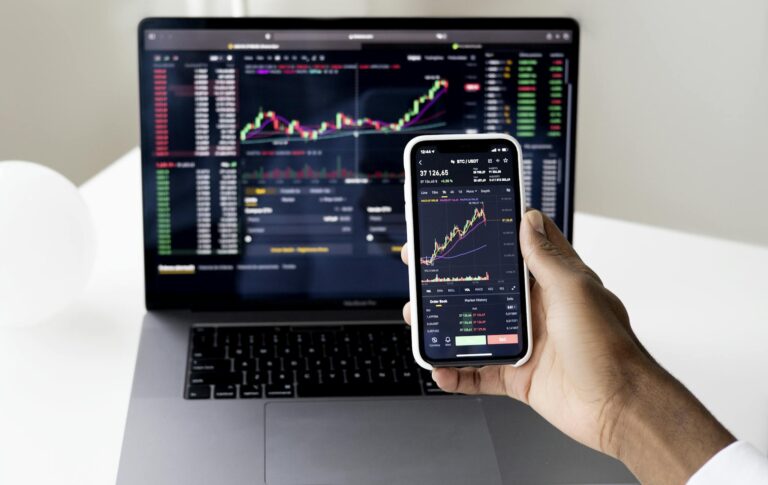 Person checking stock market graphs on laptop and smartphone, focusing on financial data.